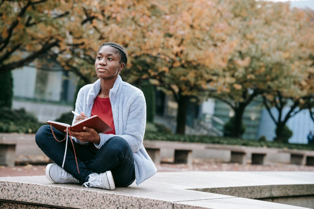Young contemplative African American woman with agenda sitting with crossed legs against autumn trees while looking forward in city