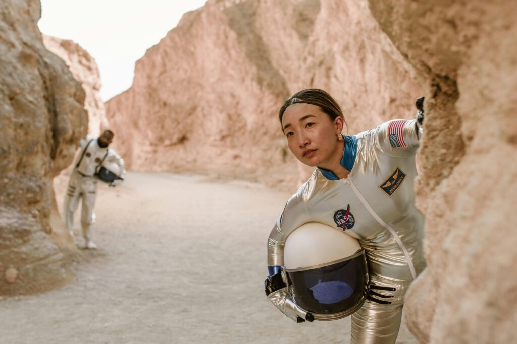 Two astronauts in silver spacesuits explore a canyon-like rocky terrain, evoking a futuristic or Mars landscape.